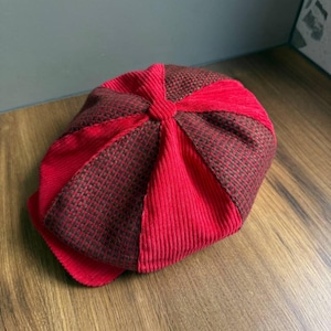 May include: A newsboy cap featuring alternating panels of solid red corduroy and a brown and black checkered fabric. The hat has a rounded crown with a central button detail and a short, stiff brim extending from the front edge.