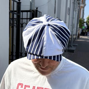 May include: A white and navy blue striped cap with a button on top. The cap has a brim and is worn by a person wearing a white t-shirt. The cap's design features alternating white and navy blue stripes.