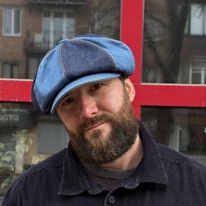 May include: A newsboy cap constructed from alternating panels of light and dark blue denim fabric. The hat features a structured brim and a button detail at the center top, showcasing visible stitching along the seams of the multi-paneled design.