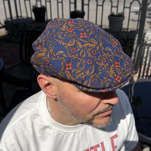 Pode incluir: Uma boina azul com um padrão floral ornamentado em laranja e dourado. A boina é usada por uma pessoa que veste uma camiseta branca com a palavra "SEATTLE" impressa em vermelho.