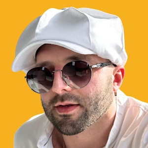 Puede incluir: Un hombre con una gorra de golf blanca y gafas redondas. Las gafas tienen un marco plateado y lentes oscuros. El hombre tiene barba y mira hacia un lado.