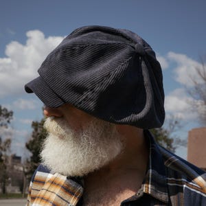 May include: A man with a long white beard is wearing a navy blue corduroy newsboy cap. He is wearing a plaid shirt with a blue, white, and orange pattern.