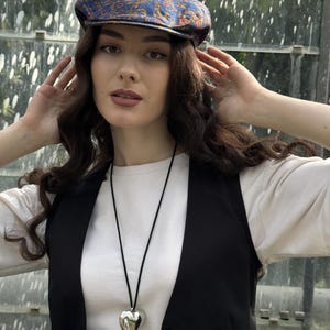 May include: A blue flat cap with an orange and gold paisley pattern, a white long-sleeve shirt, a black waistcoat, and a silver heart-shaped pendant on a thin black cord necklace.