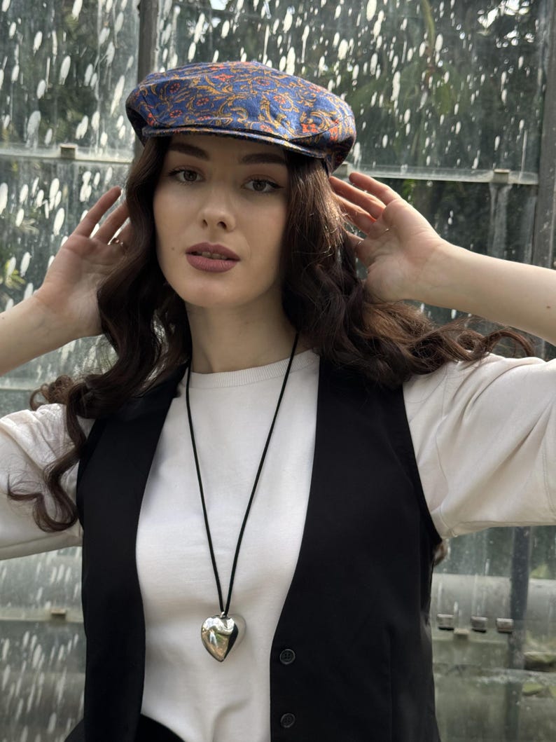 May include: A blue flat cap with an orange and gold paisley pattern, a white long-sleeve shirt, a black waistcoat, and a silver heart-shaped pendant on a thin black cord necklace.
