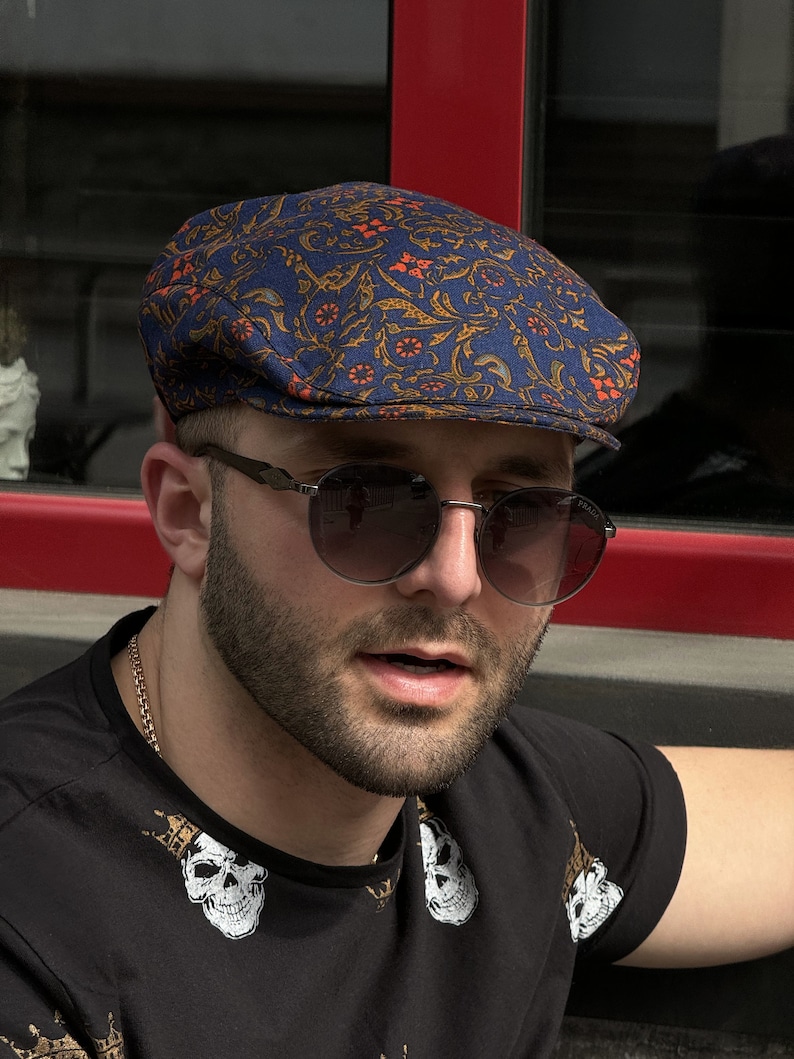 May include: A flat cap featuring a blue base with an intricate orange and gold floral pattern. The cap has a structured, rounded crown and a short, stiff brim. The fabric displays a repeating botanical design with swirling vines and small flowers.