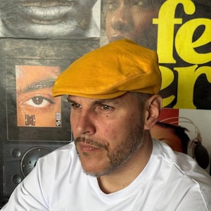Puede incluir: Una gorra plana de color amarillo mostaza sólido con una corona redondeada y una visera corta y rígida. La gorra está confeccionada con una tela lisa y mate con costuras visibles a lo largo de los bordes, manteniendo una forma estructurada.