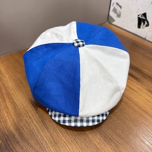 May include: A newsboy cap featuring alternating panels of solid royal blue and off-white fabric. The cap has a small, centered fabric-covered button at the top and a short, curved brim decorated with a navy blue and white gingham check pattern.
