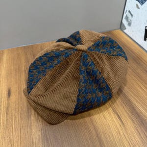 Pode incluir: Uma boina estilo newsboy construída com painéis alternados de tecido de veludo cotelê castanho e um tecido com padrão pied-de-poule azul e castanho. O chapéu apresenta um botão central na coroa e uma aba estruturada, com design texturizado.