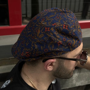 May include: A flat cap featuring a dark blue base with an intricate, repeating pattern of orange and gold floral and vine motifs. The cap has a structured, rounded crown and a short, stiff brim, showcasing a detailed textile design throughout.