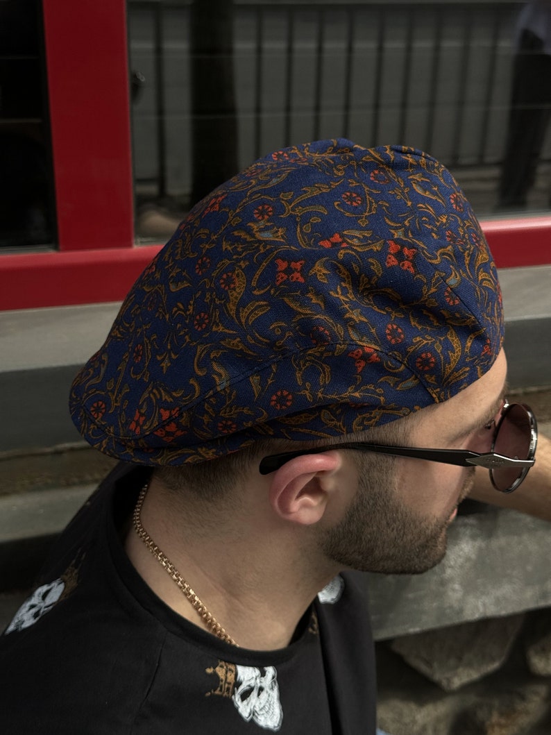 May include: A flat cap featuring a dark blue base with an intricate, repeating pattern of orange and gold floral and vine motifs. The cap has a structured, rounded crown and a short, stiff brim, showcasing a detailed textile design throughout.