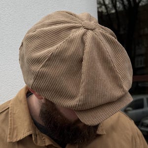 Op de afbeelding: Een beige corduroy bakkerspet met een achtdelige constructie, voorzien van een centrale met stof beklede knoop op de kruin en een korte, verstevigde klep. De pet is gemaakt van een gestructureerde stof met verticale ribbels in een lichtbruine tint.