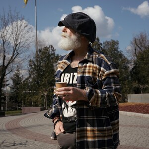 May include: A man with a white beard and a black and yellow plaid shirt is holding a brown paper cup. He is wearing a black cap and looking to the right. A yellow and blue flag is in the background.