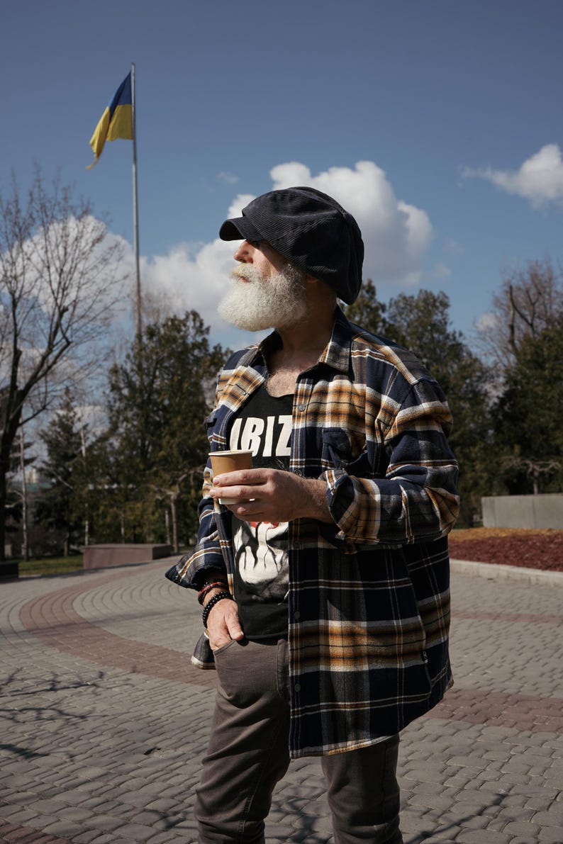 May include: A man with a white beard and a black and yellow plaid shirt is holding a brown paper cup. He is wearing a black cap and looking to the right. A yellow and blue flag is in the background.