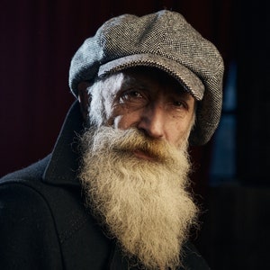Op de afbeelding: Een close-up portret van een oudere man met een lange witte baard. Hij draagt een grijze platte pet en een donkerblauw jack.