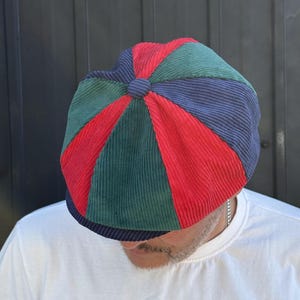 Peut inclure: Une casquette gavroche en velours côtelé multicolore composée de panneaux triangulaires alternés en rouge, vert forêt et bleu marine. Le chapeau présente une calotte arrondie avec un bouton central recouvert de tissu et une visière courte et incurvée.