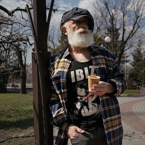 May include: A man with a white beard and a plaid shirt is standing next to a lamppost. He is holding a paper cup of coffee and wearing a black tank top with the word "IBIZ" printed on it.
