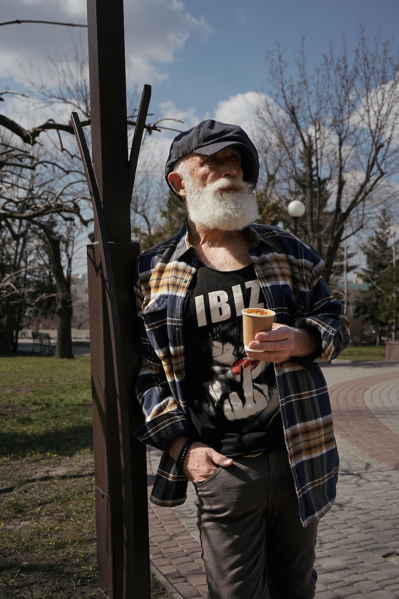May include: A man with a white beard and a plaid shirt is standing next to a lamppost. He is holding a paper cup of coffee and wearing a black tank top with the word "IBIZ" printed on it.