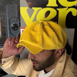 May include: A bright yellow corduroy baker boy cap featuring a multi-panelled crown, a button-topped centre, and a short, stiffened brim. The fabric has a distinct vertical ribbed texture throughout the entire surface of the hat.