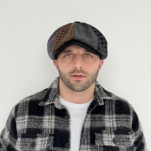 May include: A patchwork newsboy cap with a black brim. The cap features panels of brown, gray, and black patterned fabric. The person in the image is wearing a black and white plaid shirt and a white t-shirt.