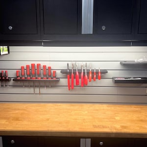 May include: A workshop wall organisation system featuring grey horizontal slat wall panels with black shelves holding various red and grey handled screwdrivers and tools, positioned above a wooden workbench with black cabinets above.