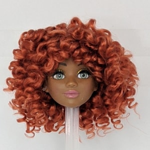 May include: A doll head with a curly, auburn red wig. The doll has brown skin, green eyes, and pink lips.