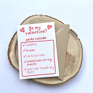May include: A Valentine's Day card with the text "Be my valentine?" and a list of perks, including "cuddles," "kisses," and "I'm hilarious." The card is white with red accents and is placed on a wooden slice with a brown envelope.
