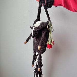 Puede incluir: Un bolso de ganchillo hecho a mano con forma de mofeta, sostenido por una persona. El bolso es principalmente negro con una raya blanca y una cola a rayas blancas y negras. Se adjunta una pequeña bolsa de fresas rojas.