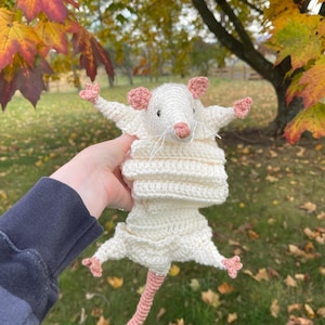K&ouml;nnte beinhalten: Ein handgefertigtes Ratten-Spielzeug aus H&auml;kelarbeit in Off-White mit rosa Akzenten. Die Ratte hat ausgestreckte Arme und einen langen Schwanz. Ohren und F&uuml;&szlig;e sind rosa. Das Spielzeug wird vor einem Hintergrund aus Herbstlaub gehalten.