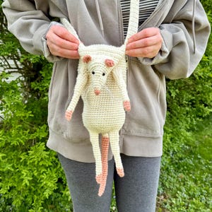 Puede incluir: Un bolso bandolera con forma de rata de ganchillo color crema con detalles en rosa en las orejas, patas y cola. El bolso tiene una correa larga y una cara detallada con ojos negros y bigotes.
