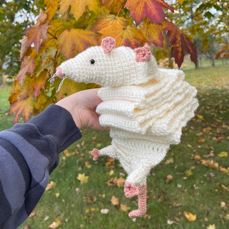 K&ouml;nnte beinhalten: Eine handgefertigte, cremefarbene H&auml;kelratte mit rosa Ohren, Nase und F&uuml;&szlig;en. Die Ratte hat einen langen, ger&uuml;schten K&ouml;rper und eine spitze Schnauze. Der Hintergrund zeigt bunte Herbstbl&auml;tter.