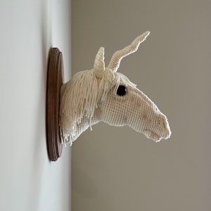 May include: A handcrafted, off-white crochet unicorn head mounted on a dark brown wooden plaque. The unicorn features a long, flowing mane, a single horn, and a dark eye. The artwork is displayed on a light-colored wall.