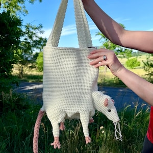 May include: A cream-colored crochet tote bag shaped like an opossum. The bag features a long pink tail, four legs with pink feet, a pointed snout with a black eye, and small pink ears. The body of the bag is textured with a dense stitch pattern.