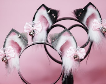 May include: Three pairs of black and white cat ears with pink fur trim and pink bows with bells. The ears are displayed on black stands.