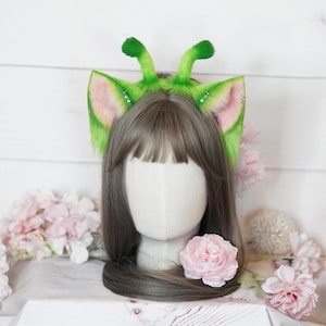 May include: A whimsical green and pink alien-themed headband with fluffy ears and antennae. The ears have pink inner details and are attached to a green headband. The antennae are also green and fluffy. The headband is displayed on a mannequin head with brown hair.