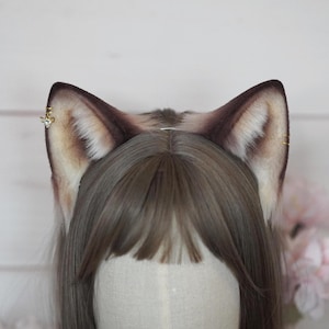 Op de afbeelding: Een paar bruine en crèmekleurige nepbont kattenoren met goudkleurige decoratieve accenten. De oren zijn bevestigd aan een hoofdband en worden op het hoofd van een persoon gedragen. De oren hebben een realistisch uiterlijk.
