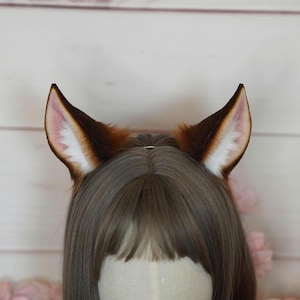 May include: A pair of brown faux fur animal ears with white tips. The ears are worn on a person's head.