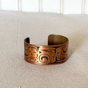 Puede incluir: Un brazalete de cobre con un diseño geométrico. El diseño es de color marrón oscuro y parece estar inspirado en el arte nativo americano.