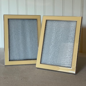 May include: Two rectangular picture frames with a light yellow border and a silver interior. The frames are leaning against a white paneled wall. The frames are empty and ready for photos.