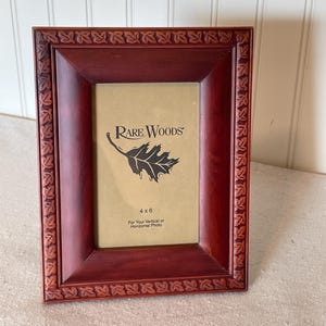Może przedstawiać: Prostokątna, ciemnoczerwona drewniana ramka na zdjęcia z dekoracyjnym wzorem liści na krawędziach. Ramka mieści zdjęcie 10 x 15 cm i zawiera logo "Rare Woods" oraz grafikę liścia.