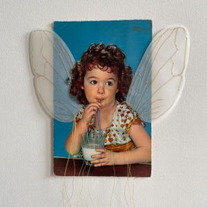 May include: A vintage photograph of a young girl with curly brown hair, drinking from a glass of milk with a blue straw. She is wearing a floral print dress and has fairy wings attached to the photo. The background is a blue colour.