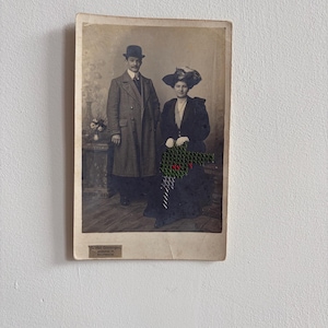 Op de afbeelding: Een vintage sepia-foto met een persoon in een lange jas en bolhoed naast een zittende persoon in een donkere jurk en hoed. Een groen en zwart kruissteekpatroon is over het midden van de afbeelding geborduurd.