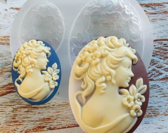 May include: Two silicone molds for making cameo-style jewelry. The molds are oval shaped and feature a raised design of a woman's profile with flowers.