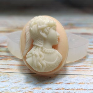 May include: Cream-coloured cameo of a woman's profile, set against a peach-coloured oval. The woman has detailed hair and jewellery. The cameo is in a clear, oval-shaped mould, likely for resin or other crafts.