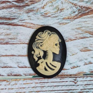 Op de afbeelding: Een zwarte cameo met een wit schedel- en skeletontwerp. De schedel heeft een vrouwelijk kapsel en kijkt naar rechts. De cameo is ovaalvormig en heeft een zwarte achtergrond.