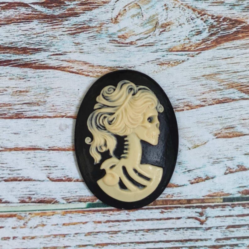Op de afbeelding: Een zwarte cameo met een wit schedel- en skeletontwerp. De schedel heeft een vrouwelijk kapsel en kijkt naar rechts. De cameo is ovaalvormig en heeft een zwarte achtergrond.