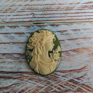 May include: A white cameo with a detailed carving of a woman's profile, surrounded by flowers. The cameo is set in a green oval-shaped frame.