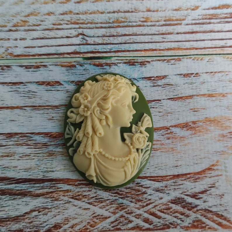 May include: A white cameo with a detailed carving of a woman's profile, surrounded by flowers. The cameo is set in a green oval-shaped frame.