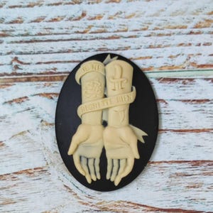 Op de afbeelding: Zwarte cameo met een wit ontwerp van twee armen met tatoeages. De tatoeages omvatten een roos, een dolk en de tekst "Je ne regrette rien".