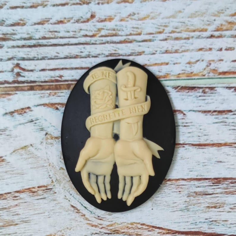 Op de afbeelding: Zwarte cameo met een wit ontwerp van twee armen met tatoeages. De tatoeages omvatten een roos, een dolk en de tekst "Je ne regrette rien".