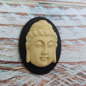 May include: A cream-colored ceramic bust of a Buddha with closed eyes, set against a black oval background.